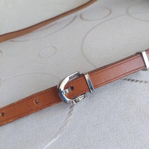May include: A brown leather belt with a silver-colored buckle. The belt is thin and appears to be made of genuine leather. The buckle is a simple, classic design. The belt is shown against a neutral background.