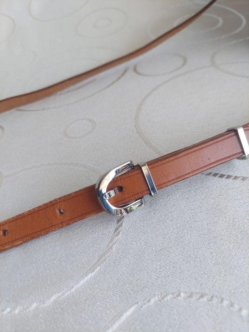 May include: A brown leather belt with a silver-colored buckle. The belt is thin and appears to be made of genuine leather. The buckle is a simple, classic design. The belt is shown against a neutral background.