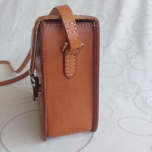 May include: A rectangular, light brown leather crossbody bag with a shoulder strap. The bag has a flap closure and visible white stitching details. The strap is attached with a metal buckle. The bag is designed for carrying essentials.