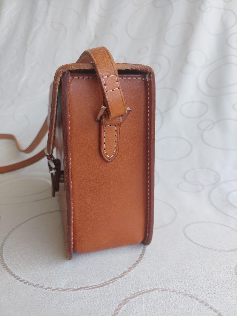 May include: A rectangular, light brown leather crossbody bag with a shoulder strap. The bag has a flap closure and visible white stitching details. The strap is attached with a metal buckle. The bag is designed for carrying essentials.