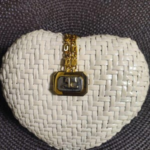 May include: A gold-toned chain necklace featuring a rectangular pendant with a polished gold border and a silver-toned center. The center of the pendant displays the letters AC in a stylized font. The chain consists of repeating interlocking links.