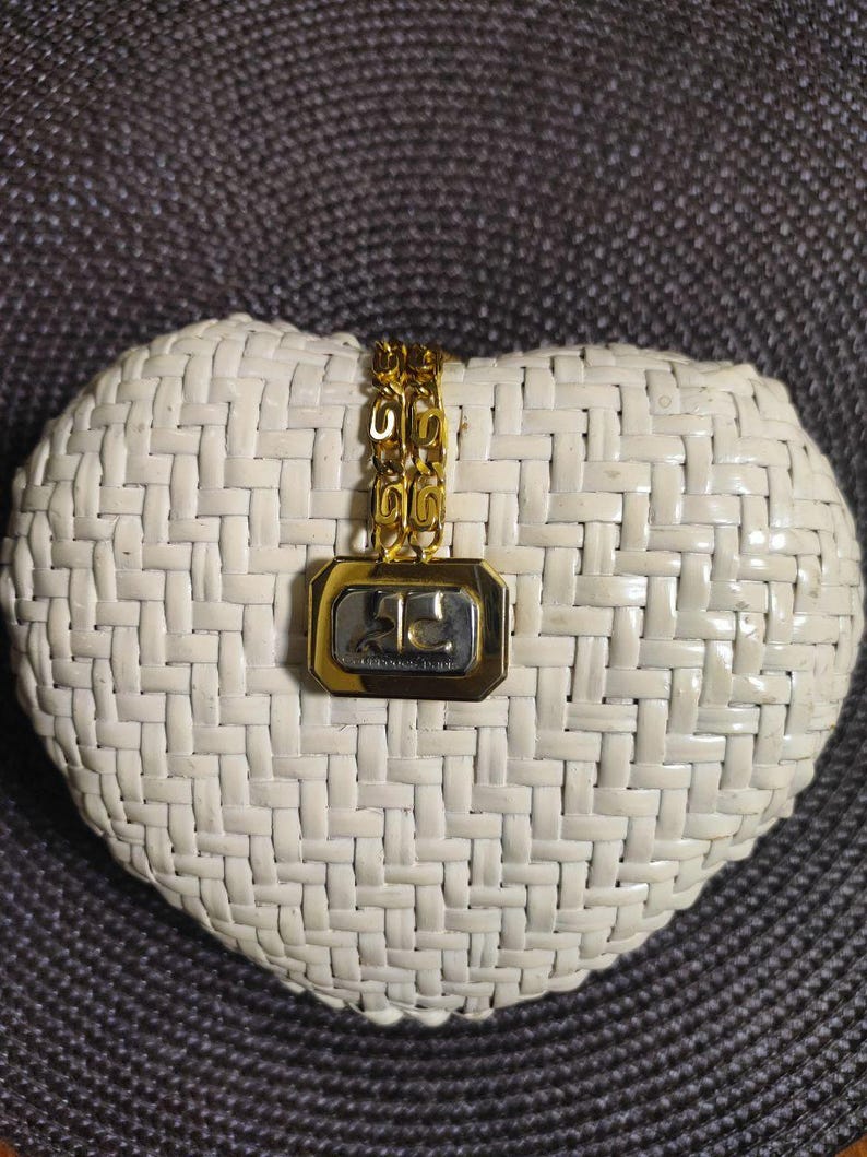 May include: A gold-toned chain necklace featuring a rectangular pendant with a polished gold border and a silver-toned center. The center of the pendant displays the letters AC in a stylized font. The chain consists of repeating interlocking links.