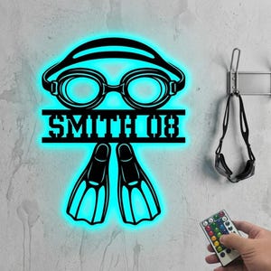 Può includere: Un'opera d'arte da parete in metallo nero con una luce blu neon, raffigurante occhialini da nuoto, pinne e il nome "SMITH 08". Un telecomando è tenuto in mano. Un paio di occhialini da nuoto sono appesi al muro.