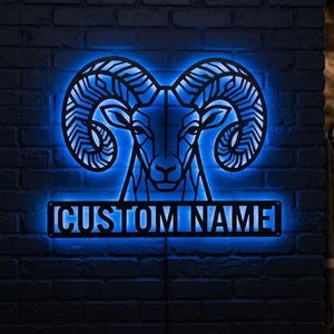 Puede incluir: Un letrero de pared de metal negro con la silueta geométrica de una cabeza de carnero con grandes cuernos curvos, iluminado por una luz LED azul brillante. Debajo del carnero, una placa rectangular muestra el texto CUSTOM NAME en mayúsculas.
