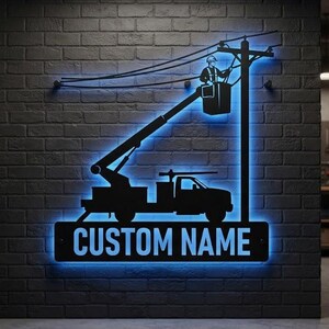 May include: A black metal wall sign featuring a silhouette of a utility truck with an extended boom lift and a worker repairing power lines. The sign is illuminated by a bright blue LED light and includes a rectangular base with the text CUSTOM NAME.