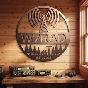 Pode incluir: Uma placa circular de metal para parede com uma torre de rádio com ondas de sinal no topo, o texto W7RAD no centro e uma silhueta de uma casa e pinheiros na parte inferior com o texto EST. 2025, com acabamento em tom de bronze escuro.