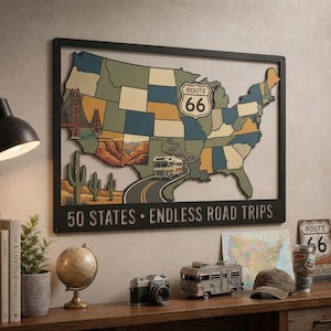 Peut inclure: Une décoration murale rectangulaire en métal noir représentant une carte multicolore des États-Unis. La carte inclut un panneau Route 66, un camping-car sur une route sinueuse, un paysage de cactus et le Golden Gate Bridge. Le texte indique 50 STATES • ENDLESS ROAD TRIPS.