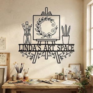 May include: A black metal wall sign featuring an easel with a canvas displaying a floral wreath, a jar of paintbrushes, and a cup of pencils. The text LINDA'S ART SPACE is centered below the easel, accented by decorative vine flourishes.