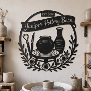 May include: A circular black metal wall sign featuring the text Juniper Pottery Barn and EST. 2016. The design includes a central pottery wheel with a bowl, a vase, and sculpting tools, all framed by a decorative border of leaves and flowers.