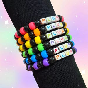 Könnte beinhalten: Ein Stapel bunter Perlenarmbänder. Jedes Armband zeigt das Wort "PLUR" mit Buchstabenperlen. Die Armbänder sind in einem Regenbogen aus Farben, darunter Rosa, Orange, Gelb, Grün, Blau und Lila, mit schwarzen Perlen.