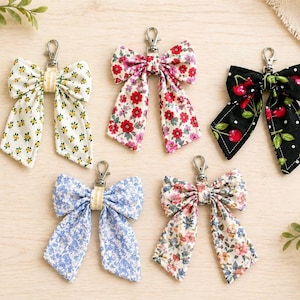 May include: Five fabric bow keychains with metal lobster clasps. The top row features a white bow with yellow flowers, a pink floral bow, and a black bow with a red cherry print. The bottom row includes a blue floral bow and a multi-coloured floral bow.
