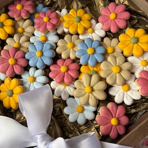 Op de afbeelding: Een verzameling bloemvormige koekjes met gekleurd glazuur in tinten roze, geel, lichtblauw en beige, gerangschikt in een houten doos bekleed met goudkleurig vloeipapier en onderaan vastgebonden met een witte strik.