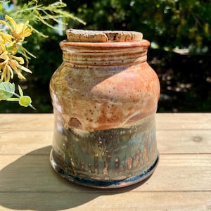 May include: A handmade ceramic jar with a cork lid. The jar features a color gradient, transitioning from a reddish-brown top to a teal-green base. The jar is approximately 5 inches tall and sits on a wooden surface.