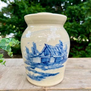 May include: A cream-colored ceramic jar with a wide rim and a lid. The exterior features a hand-painted blue ink illustration of a barn surrounded by trees and foliage. The design includes small birds in the sky above the structure.