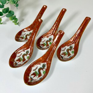 May include: A set of four ceramic soup spoons featuring a white interior with a painted dragon motif in green, brown, and gold. The exterior of each spoon is finished in a deep red glaze with a gold crackle pattern throughout the handle.
