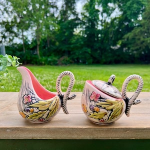 May include: A pink and white ceramic creamer and sugar bowl set. The creamer has a curved spout and a pink interior. The sugar bowl has a lid. Both pieces feature floral and leaf designs with black accents and looped handles.