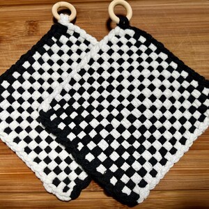 May include: Two square, woven potholders with a black and white checkerboard pattern. Each potholder has a white border and a wooden ring for hanging. The potholders are on a wooden cutting board.