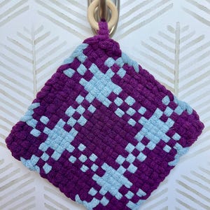 May include: A square, woven potholder in shades of purple and light blue. The potholder features a geometric pattern and a wooden ring for hanging. The design is a mix of dark purple and light blue squares, creating a textured appearance.