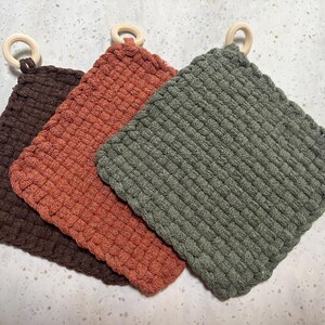 May include: Three square, woven potholders in shades of brown, orange, and green. Each potholder has a wooden ring attached for hanging. The potholders are made of a thick, textured material.
