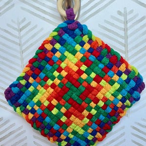 May include: A square, woven hot pad in a vibrant rainbow of colors, including red, orange, yellow, green, blue, and purple. It has a loop made of purple yarn and a wooden ring for hanging. The hot pad is approximately 7 inches square.