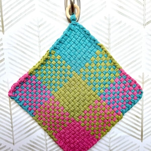 May include: A colorful, woven hot pad in a diamond shape. The pad features a pattern of turquoise, pink, and olive green squares. A wooden ring is attached to the top corner for hanging. The hot pad is approximately 8 inches wide.