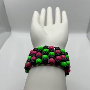 May include: A wide bracelet made of black, neon green, and pink beads. The beads are strung together to create a layered effect. The bracelet is displayed on a white hand.