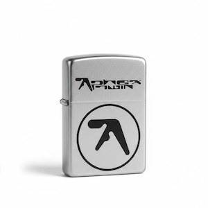 May include: A silver metal lighter with the Aphex Twin logo printed in black. The logo is on the top and bottom of the lighter. The bottom logo is a black and white circle with a stylized face.