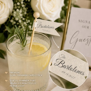 May include: A cocktail stirrer featuring a small, cream-coloured flag made of ivory shimmer cardstock. The flag is printed with the text The Bartolones and the date 09.18.2027. The stirrer is placed in a glass containing a clear liquid, ice, and a lemon slice.