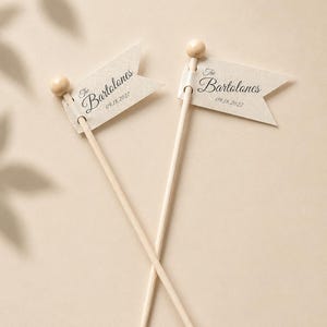 May include: Two light beige wooden sticks topped with small wooden spheres, each featuring a swallowtail flag made of textured off-white paper. The flags are printed with the text The Bartolones and the date 09.18.2027 in a dark script font.