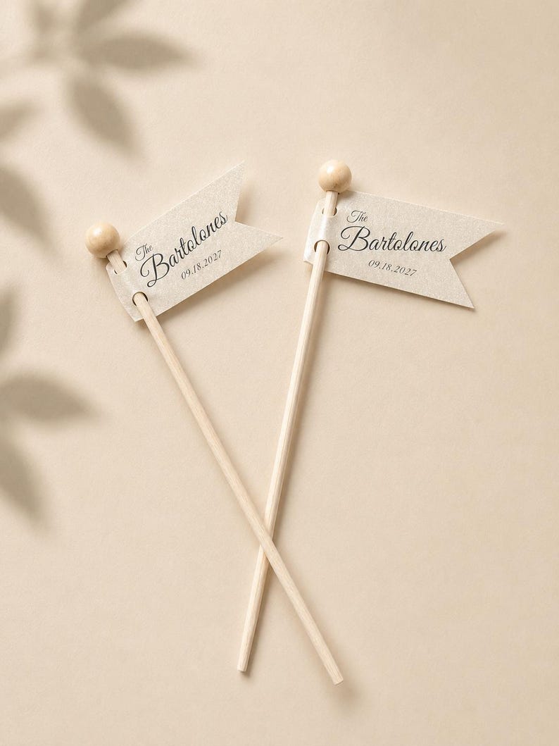 May include: Two light beige wooden sticks topped with small wooden spheres, each featuring a swallowtail flag made of textured off-white paper. The flags are printed with the text The Bartolones and the date 09.18.2027 in a dark script font.