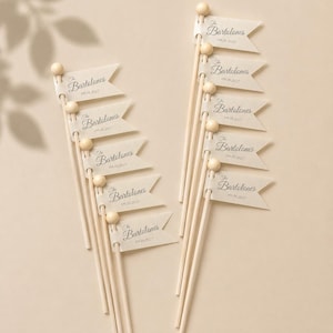 May include: A set of ten light-coloured wooden sticks, each topped with a small wooden bead and a small off-white paper flag. Each flag features the printed text The Bartolones and the date 09.18.2027 in a dark script font with a swallowtail edge.