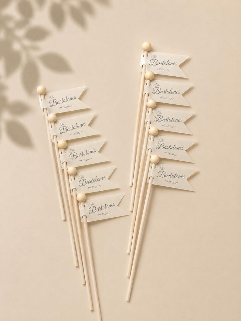 May include: A set of ten light-coloured wooden sticks, each topped with a small wooden bead and a small off-white paper flag. Each flag features the printed text The Bartolones and the date 09.18.2027 in a dark script font with a swallowtail edge.