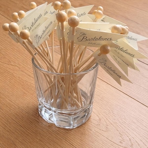 May include: A clear glass tumbler filled with wooden cocktail picks, each topped with a small round bead and a cream-coloured paper flag. The flags are printed with the text The Bartalones and the date 04.15.2027 in a dark script font.