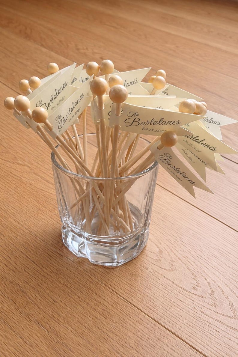 May include: A clear glass tumbler filled with wooden cocktail picks, each topped with a small round bead and a cream-coloured paper flag. The flags are printed with the text The Bartalones and the date 04.15.2027 in a dark script font.