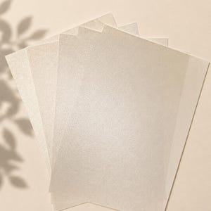 May include: A stack of rectangular sheets of paper with a shimmering, pearlescent finish. The paper has a light cream or off-white colour with a fine, reflective texture that catches the light, creating a subtle sparkle across the surface.