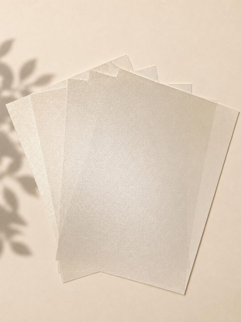 May include: A stack of rectangular sheets of paper with a shimmering, pearlescent finish. The paper has a light cream or off-white colour with a fine, reflective texture that catches the light, creating a subtle sparkle across the surface.