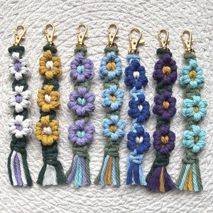 May include: A collection of handmade macrame keychains featuring colorful flower designs. Each keychain has a gold-colored clasp and a tassel at the bottom. The flowers are in various colors, including white, yellow, purple, blue, and teal.
