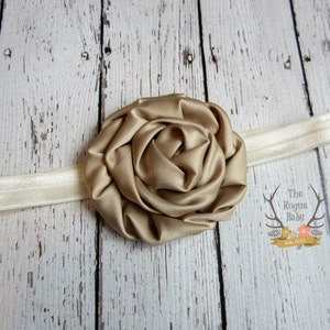 May include: A gold satin flower headband with a white elastic band. The headband is on a white wooden background.