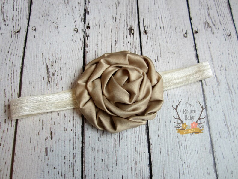 May include: A gold satin flower headband with a white elastic band. The headband is on a white wooden background.