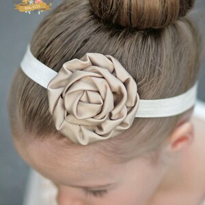 May include: A white headband with a large, beige fabric rose accent. The headband is worn by a young person with their hair in a bun.