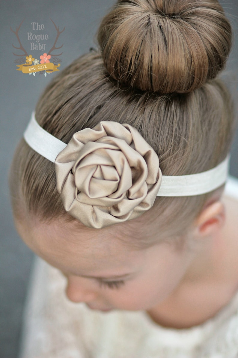 May include: A white headband with a large, beige fabric rose accent. The headband is worn by a young person with their hair in a bun.