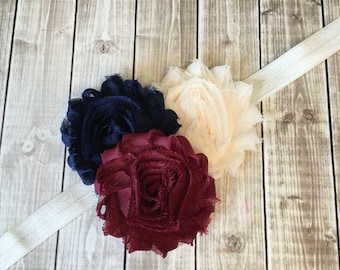 May include: A headband with three fabric flowers in navy blue, burgundy, and cream colors. The headband is made of a white elastic material.