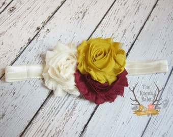 May include: A white, burgundy, and yellow flower headband. The headband is made of a white elastic band with three fabric flowers attached. The flowers are made of a soft, ruffled fabric.