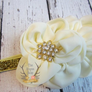 May include: A white fabric flower headband with a gold rhinestone centre. The headband is made of a sparkly gold elastic.