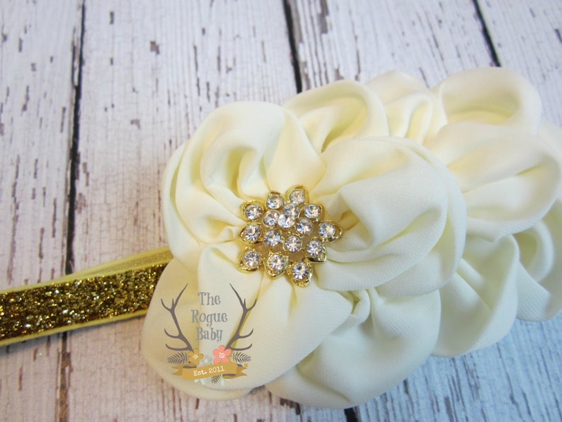May include: A white fabric flower headband with a gold rhinestone centre. The headband is made of a sparkly gold elastic.