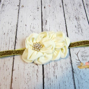 May include: A white fabric flower headband with a gold glitter elastic band and a rhinestone centre.