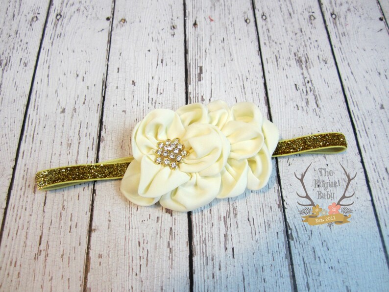 May include: A white fabric flower headband with a gold glitter elastic band and a rhinestone centre.