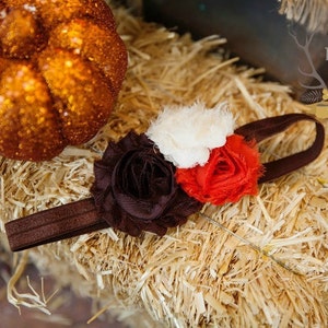 May include: A brown headband with three fabric flowers in shades of brown, white, and red. The headband is on a bed of straw with a small pumpkin in the background. The text "The Rogue Baby Est. 2011" is visible in the top right corner of the image.