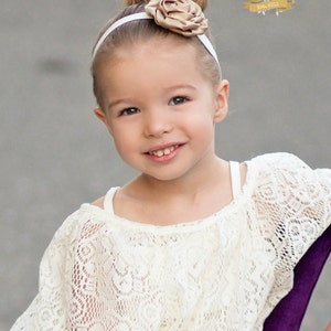 May include: A young girl with a white headband and a brown flower on top of her head is wearing a white lace top and blue jeans. She is smiling and looking at the camera.