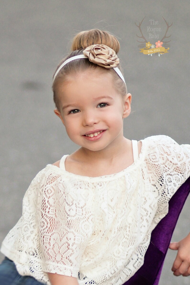 May include: A young girl with a white headband and a brown flower on top of her head is wearing a white lace top and blue jeans. She is smiling and looking at the camera.
